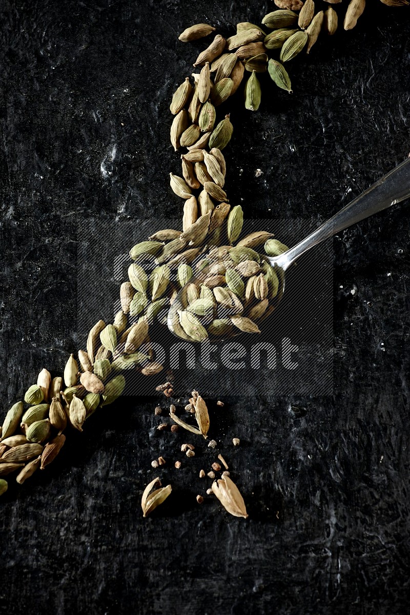 A Metal spoon full of cardamom seeds and some seeds beside it on a textured black flooring