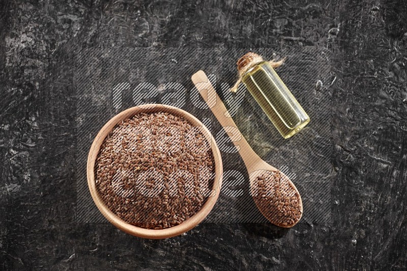 A wooden bowl and spoon full of flaxseeds and a glass bottle of flaxseeds oil on a textured black flooring