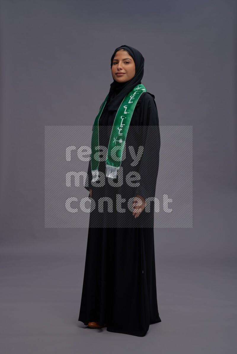 Saudi woman wearing Abaya standing holding Saudi flag on gray background