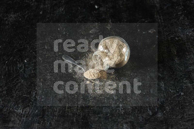 وعاء زجاجي للتوابل مقلوب وملعقة معدنية ممتلئان ببودرة الهيل والبودرة تناثرت منه على خلفية سوداء