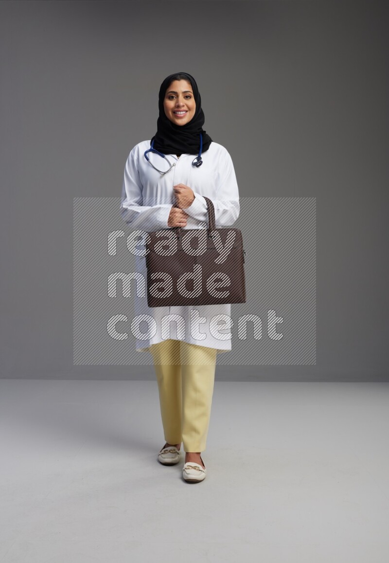 Saudi woman wearing lab coat with stethoscope standing holding bag on Gray background