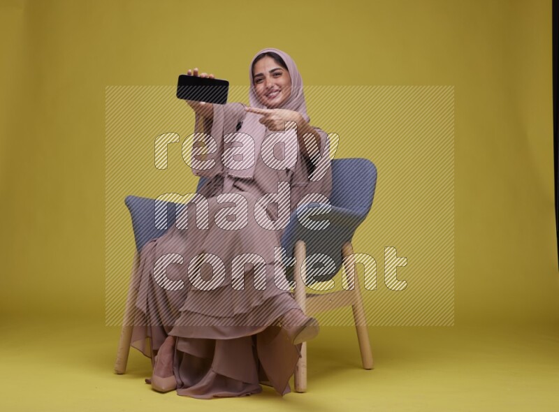 A woman Showing Her Phone Screen Setting  on a Yellow Background wearing Brown Abaya with Hijab