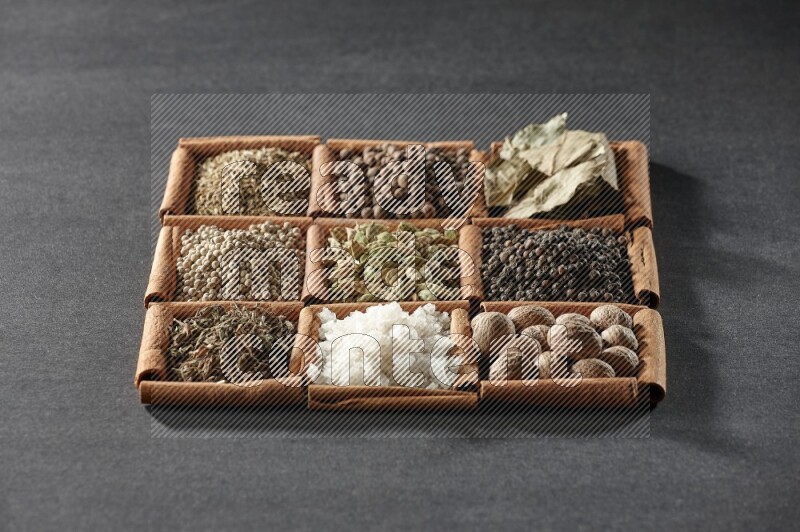 9 مربعات من أعواد القرفة ممتلئة بالهيل في المنتصف محاطة بجوزة الطيب والفلفل الأسود وورق الغار والبهارات والكمون والفلفل الأبيض والريحان المجفف والملح على أرضية سوداء