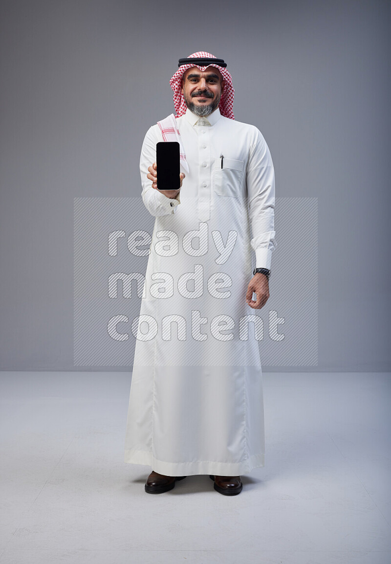 Saudi man Wearing Thob and red Shomag standing showing phone to camera on Gray background