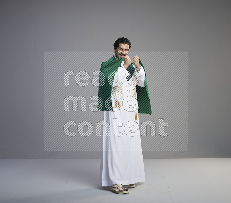 A Saudi man standing wearing thob and saudi flag scarf with face painting wrapping big Saudi flag on gray background