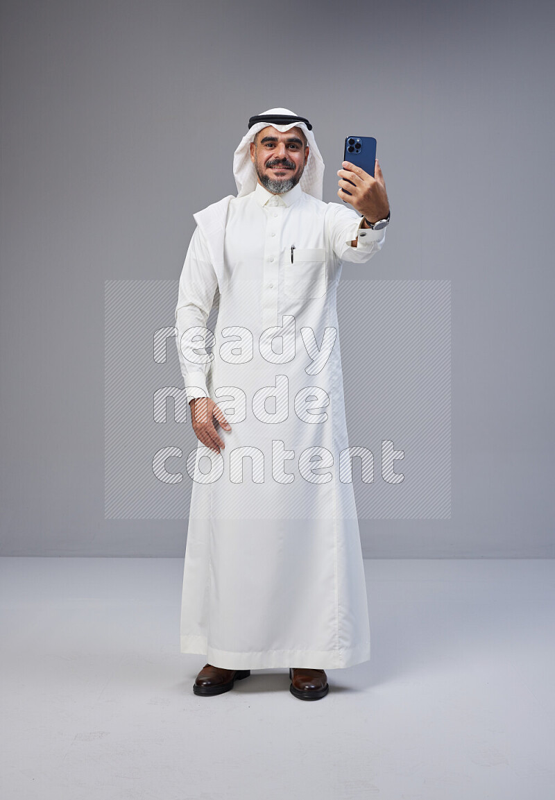 Saudi man Wearing Thob and white Shomag standing taking selfie on Gray background