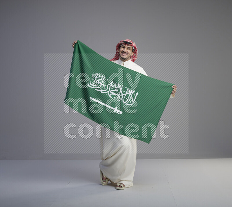 A saudi man standing wearing thob and red shomag holding big saudi flag on gray background