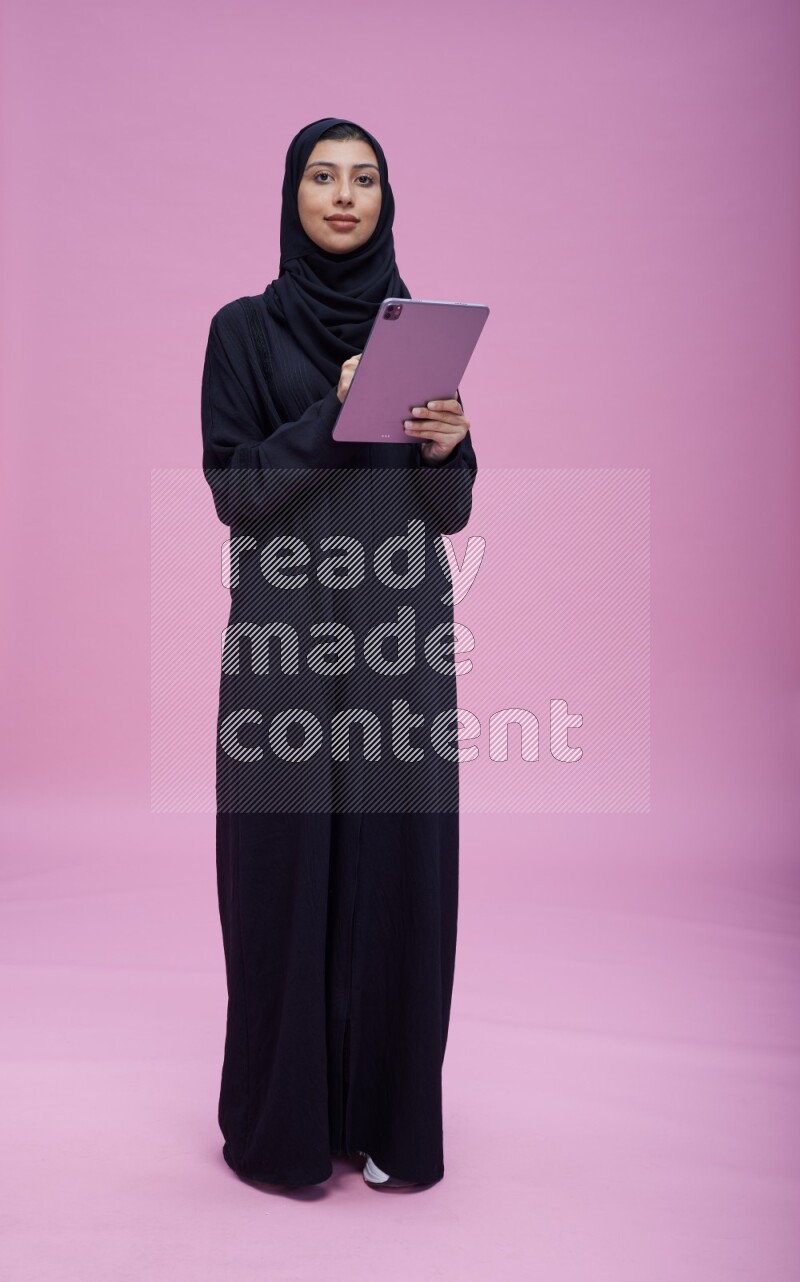 Saudi woman wearing Abaya standing working on tablet on pink background