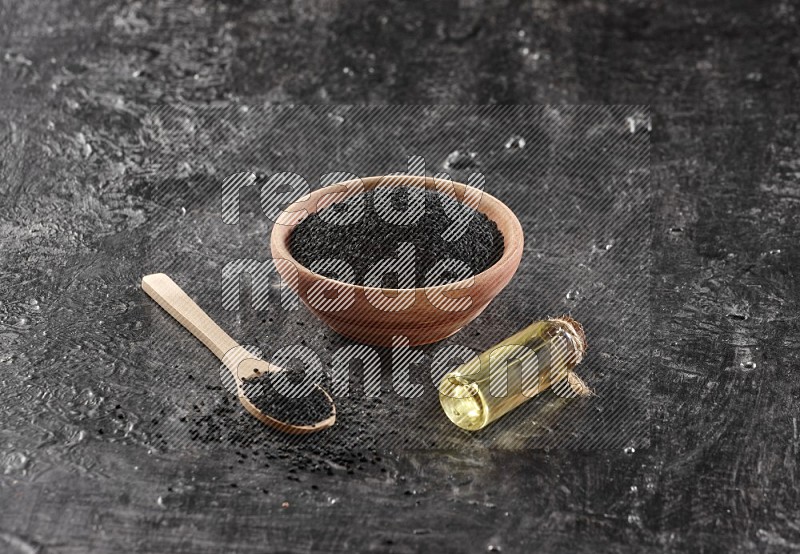 A wooden bowl and spoon full of black seeds and a bottle of black seeds oil on a textured black flooring in different angles