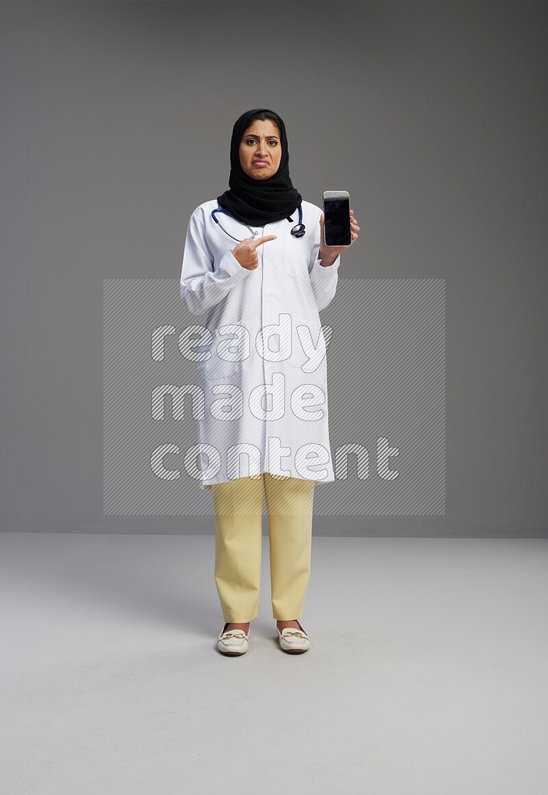 Saudi woman wearing lab coat with stethoscope standing showing phone to camera with sign in the back on Gray background