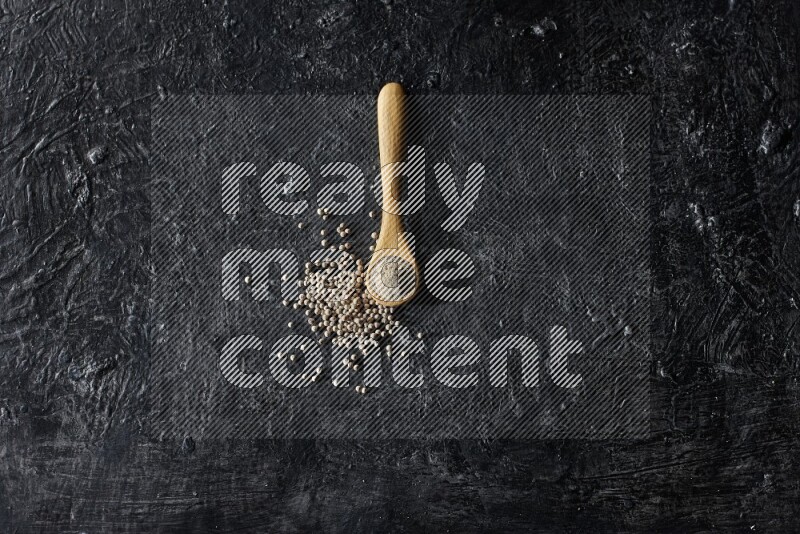 A wooden spoon full of white pepper powder with white pepper beads on textured black flooring
