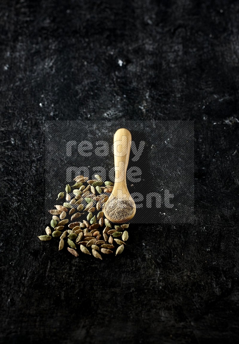 A wooden spoon full of cardamom powder and cardamom seeds next of it on textured black flooring