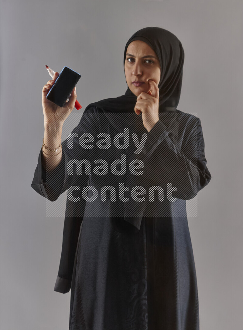 Woman in black abaya and hijab holding a marker pen to write on a transparent board with different reactions and expressions on grey background