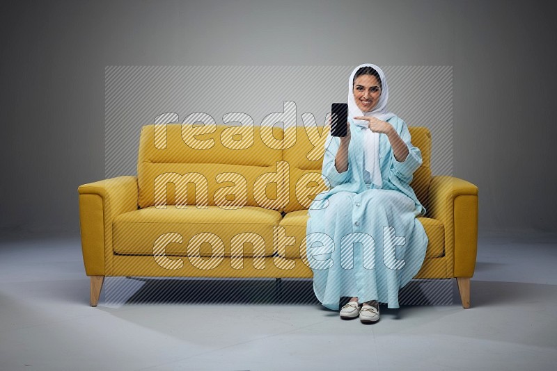 A Saudi woman wearing a light blue Abaya and white head scarf sitting on a yellow sofa and showing her phone's screen vertically and horizontally eye level on a grey background