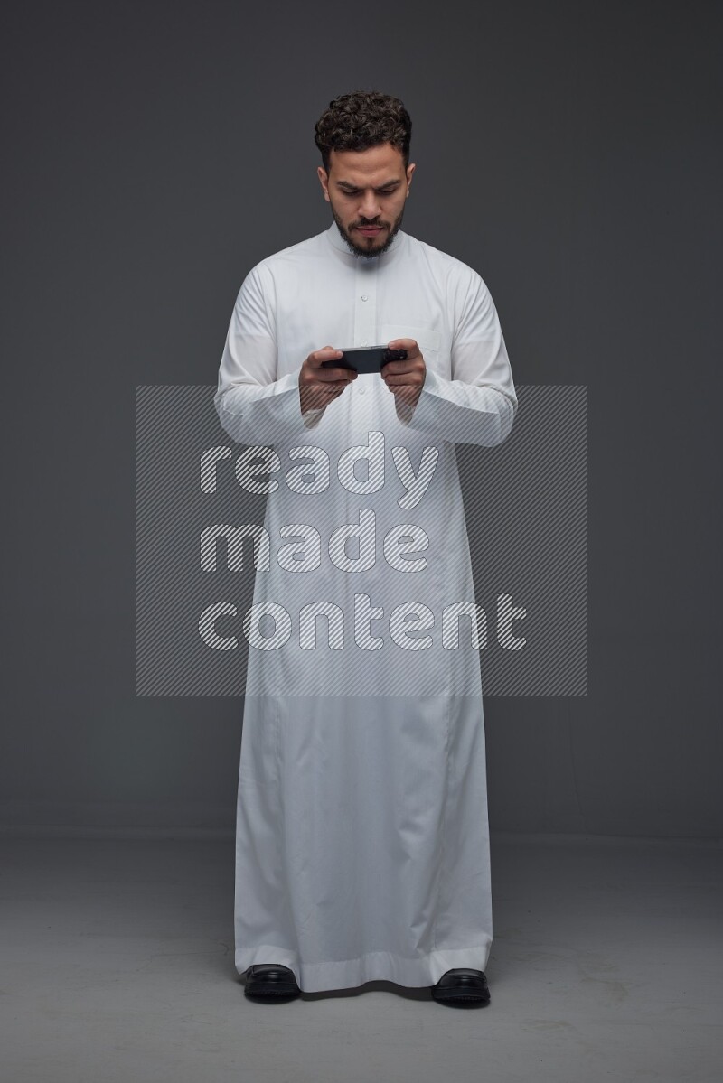 A Saudi man wearing Thobe and playing using his phone horizontally playing video games while standing and making different poses eye level on a gray background
