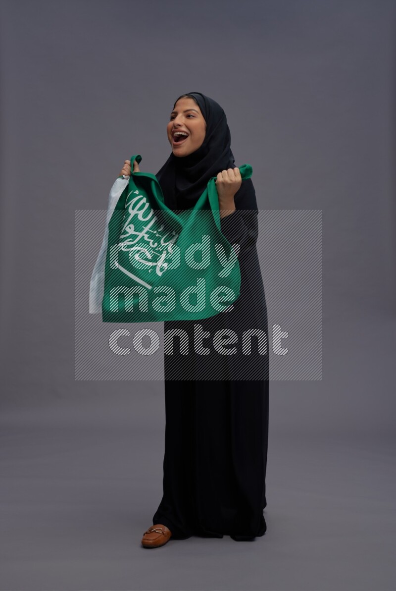 Saudi woman wearing Abaya standing holding Saudi flag on gray background
