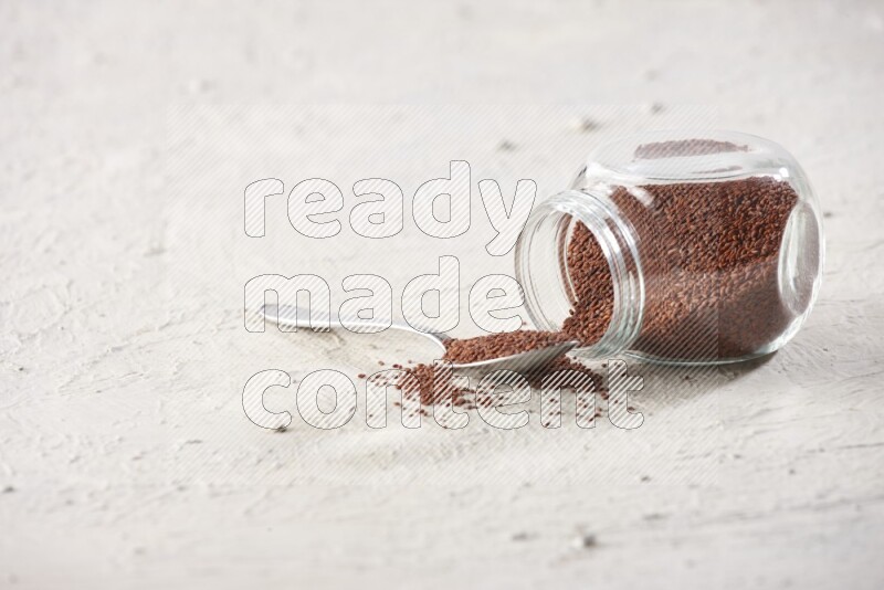 A glass spice jar and a metal spoon full of garden cress seeds and jar is flipped with fallen seeds on a textured white flooring