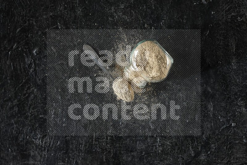 A flipped glass spice jar and metal spoon full of cardamom powder and powder spilled out of it on textured black flooring