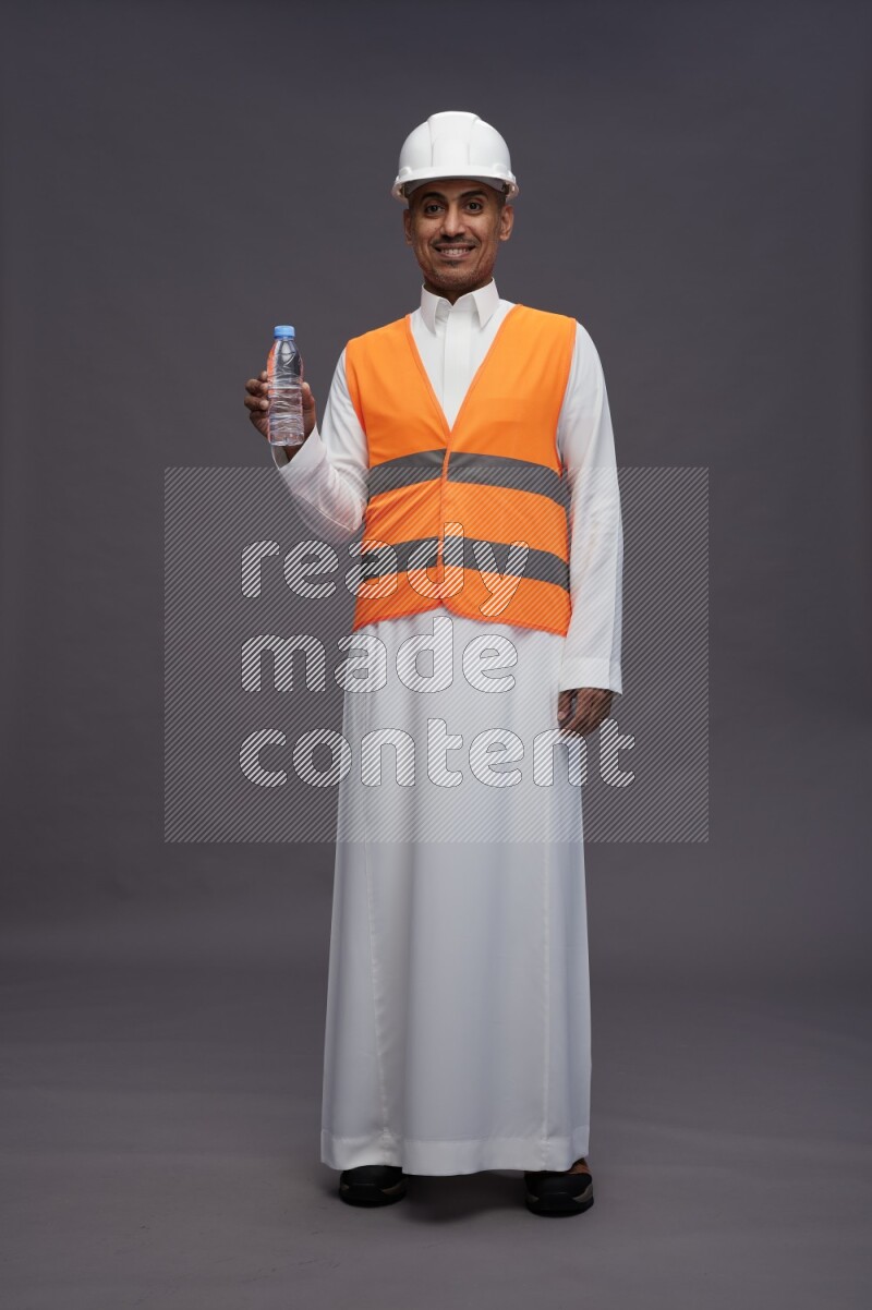 Saudi man wearing thob with engineer vest standing drinking water on gray background