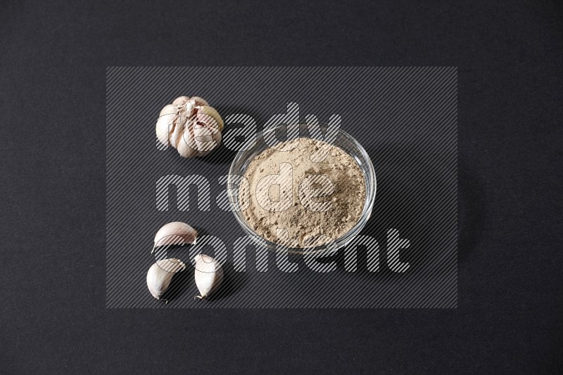 A glass bowl full of garlic powder with garlic bulb and cloves beside it on a black flooring in different angles