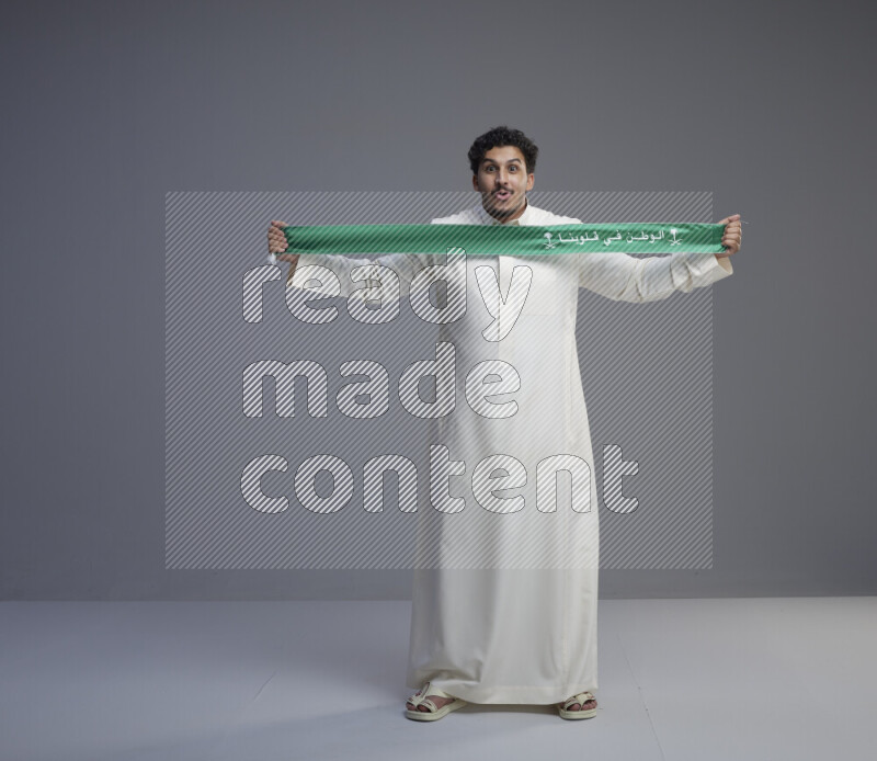 A Saudi man standing wearing thob holding Saudi flag scarf on gray background