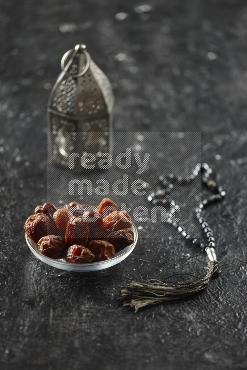 A silver lantern with different drinks, dates, nuts, prayer beads and quran on textured black background