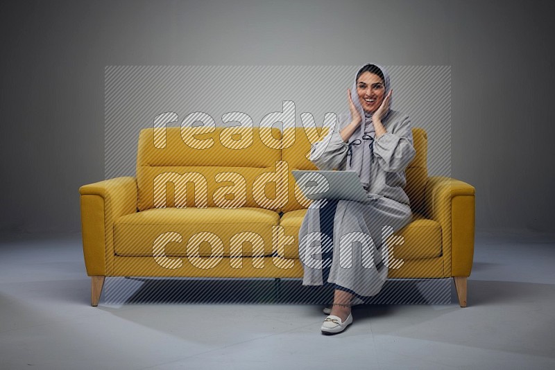 A Saudi woman wearing a light gray Abaya and head scarf sitting on a yellow sofa and using her laptop eye level on a grey background
