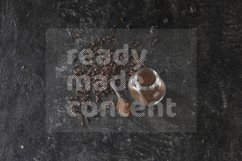 A glass spice jar and a metal spoon full of cloves powder with cloves spread on textured black flooring