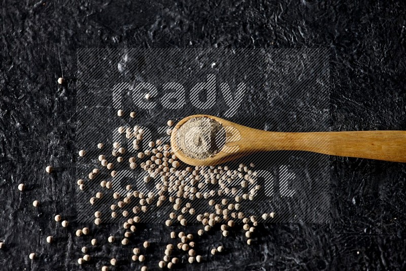 A wooden spoon full of white pepper powder with white pepper beads on textured black flooring