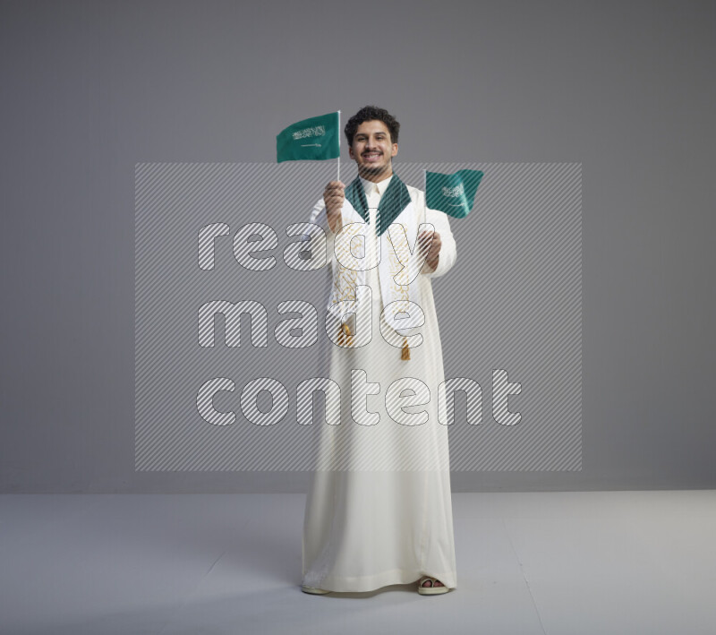 A Saudi man standing wearing thob and saudi flag scarf and holding small saudi flag on gray background
