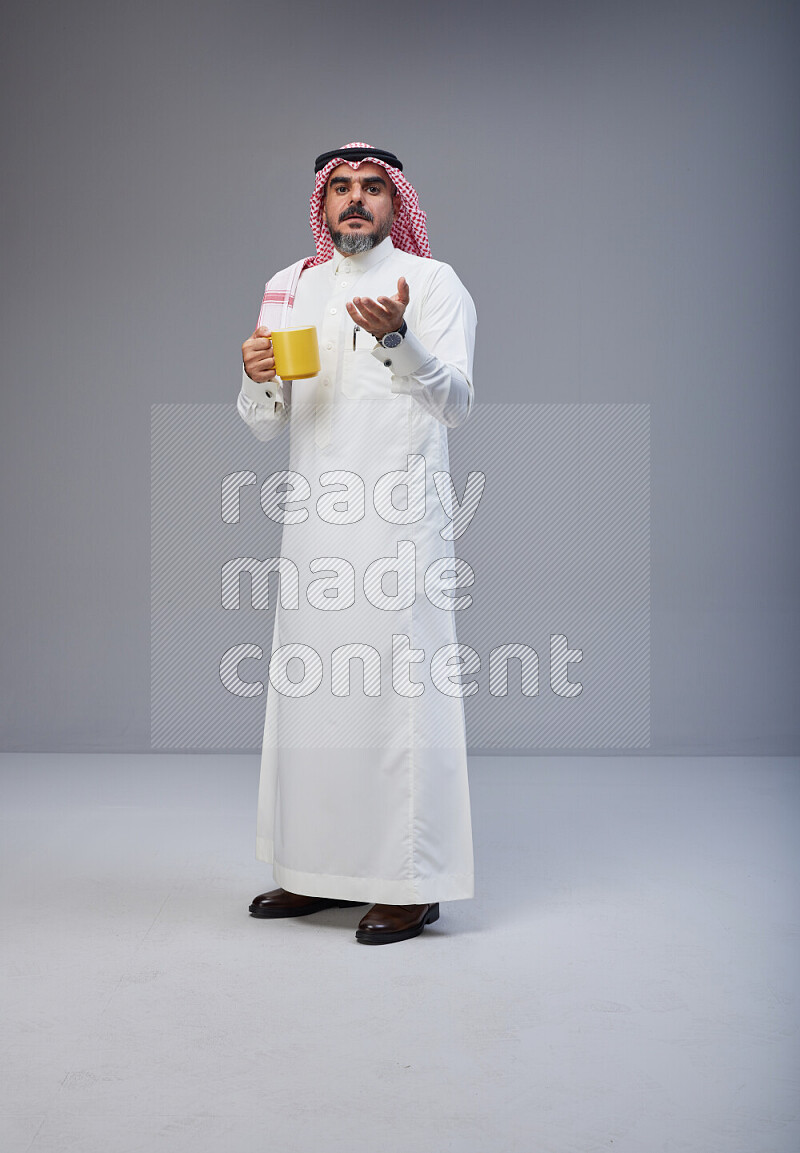 Saudi man Wearing Thob and red Shomag standing holding mug on Gray background