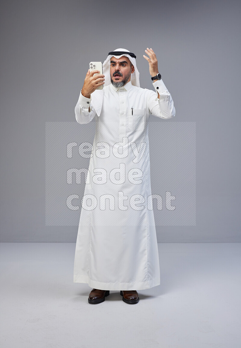 Saudi man Wearing Thob and white Shomag standing texting on phone on Gray background