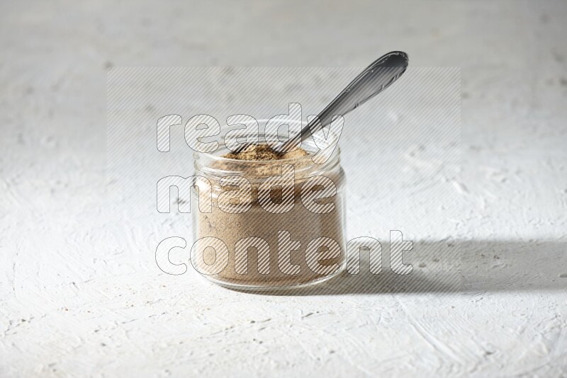A glass jar and a metal spoon full of cumin powder on textured white flooring