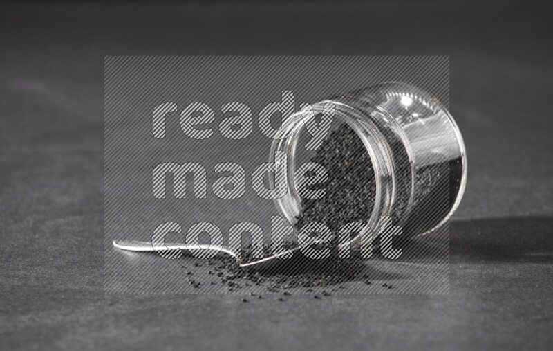 A flipped glass jar full of black seeds and seeds spread out with a metal spoon full of the seeds on a black flooring