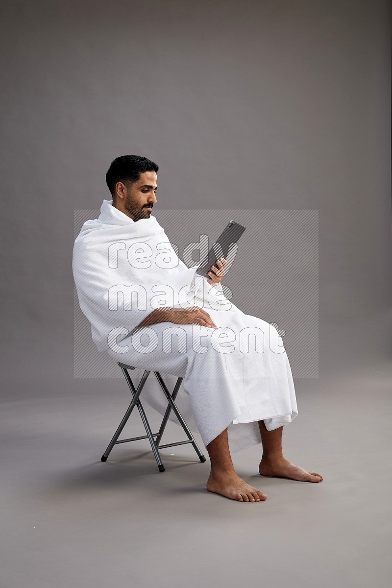 A man wearing Ehram sitting on chair holding a tablet on gray background