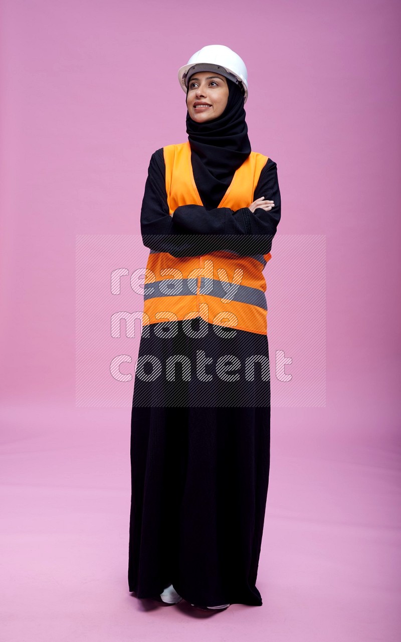 Saudi woman wearing Abaya with engineer vest and helmet standing with crossed arms on pink background