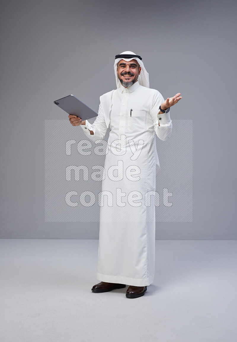 Saudi man Wearing Thob and white Shomag standing working on tablet on Gray background