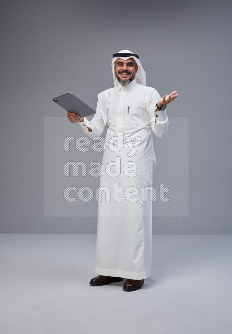 Saudi man Wearing Thob and white Shomag standing working on tablet on Gray background