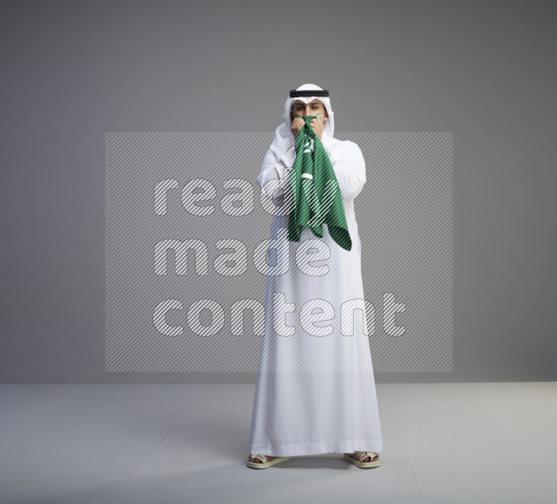 A Saudi man standing wearing thob and white shomag with face painting kissing big Saudi flag on gray background