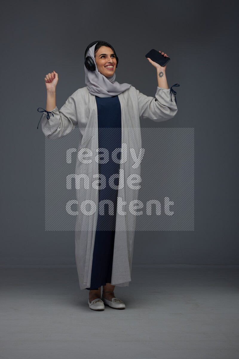 A Saudi woman wearing a light gray Abaya and head scarf standing and listening to music on a grey background