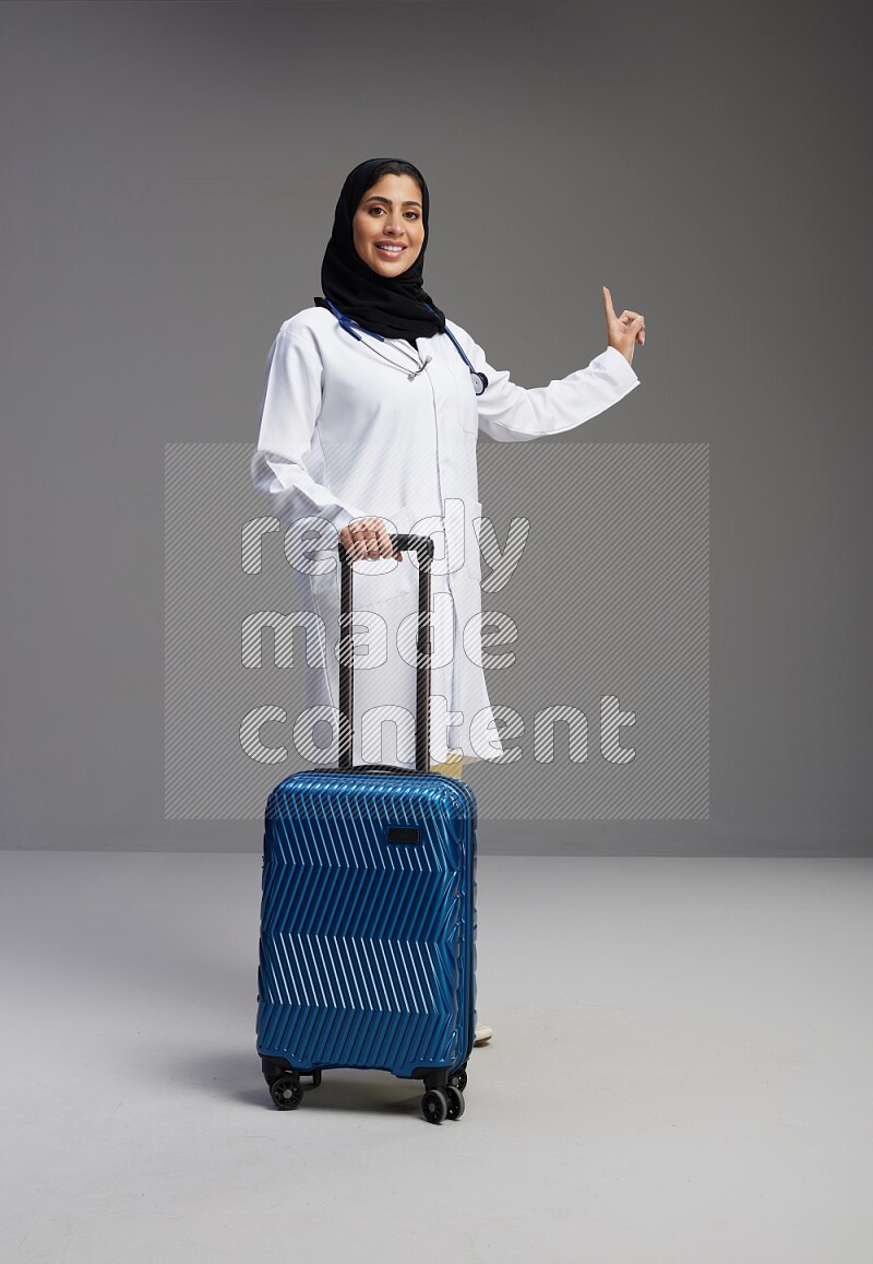 Saudi woman wearing lab coat with stethoscope standing holding Travel bag on Gray background