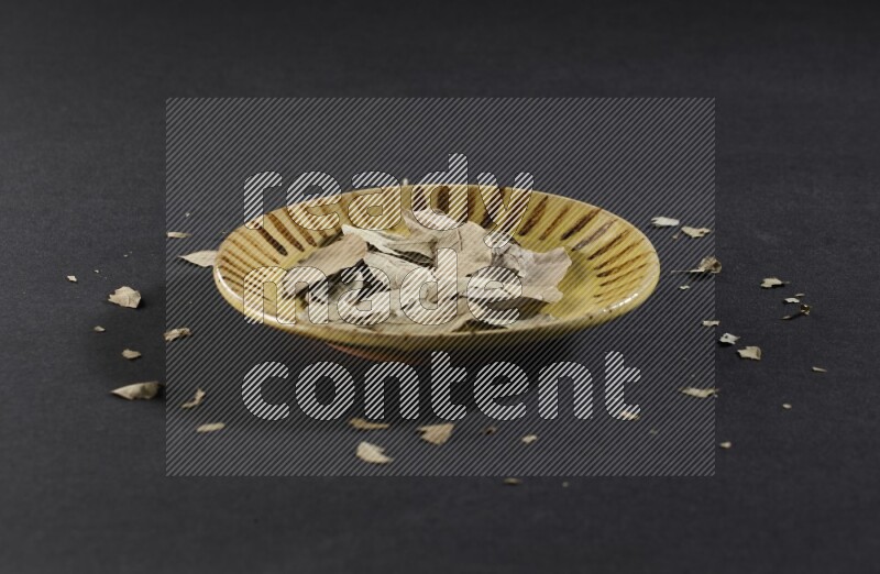 A multicolored pottery plate filled with dried bay leaves on black flooring