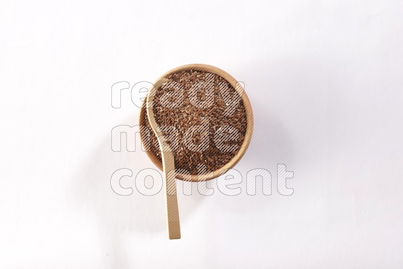 A wooden bowl full of flax seeds and a wooden spoon full of flax seeds in it on a white flooring