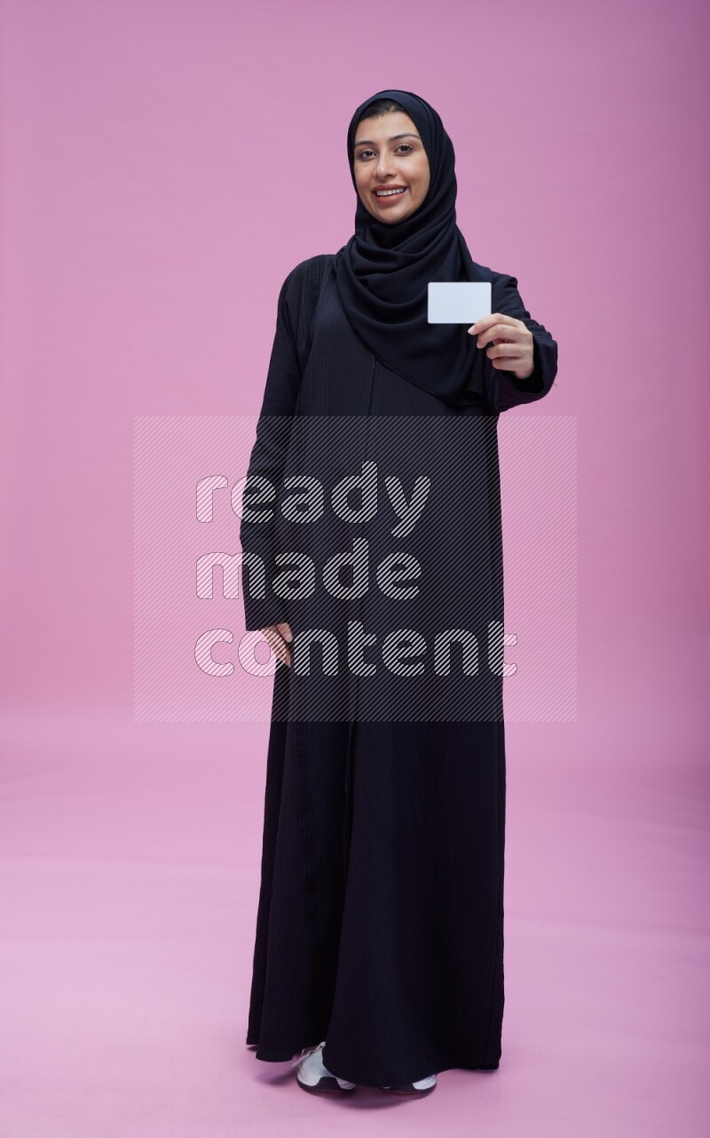 Saudi woman wearing Abaya standing holding ATM card on pink background