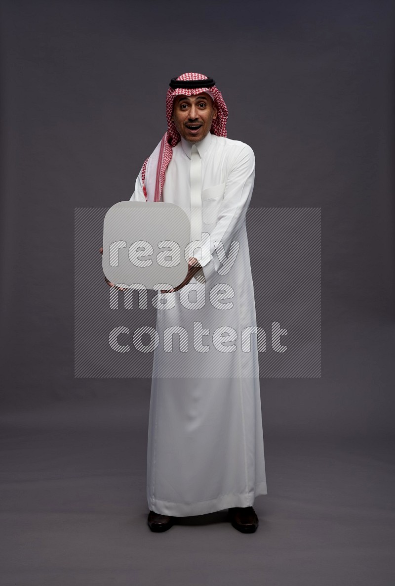 Saudi man wearing thob and shomag standing holding social media sign on gray background