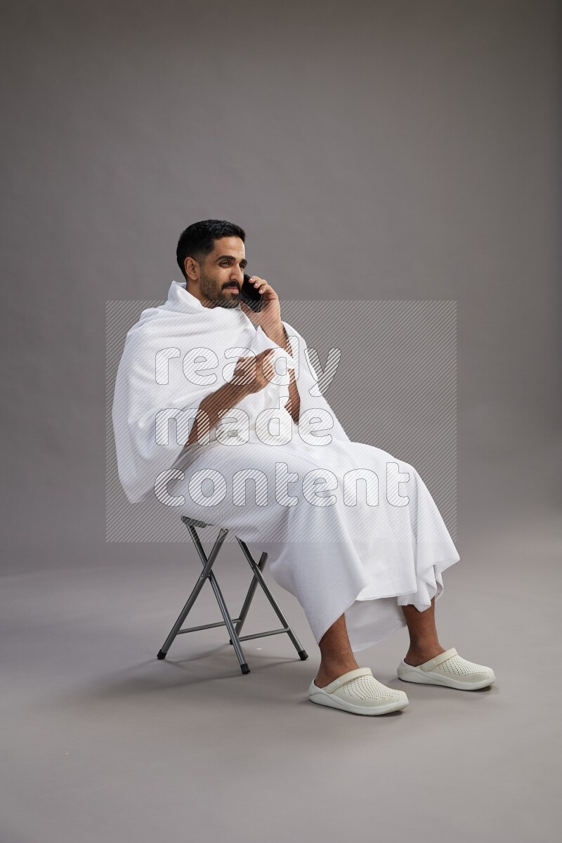 A man wearing Ehram sitting on chair talking on the phone on gray background
