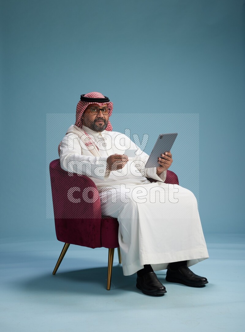 Saudi Man with shimag sitting on chair holding ATM card while working on tablet on blue background