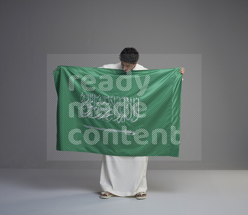 A Saudi man standing wearing thob holding big Saudi flag on gray background