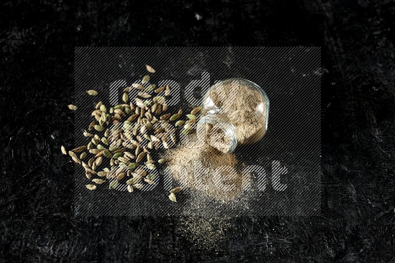 A flipped glass spice jar full of cardamom powder and powder spilled out of it with cardamom seeds spreaded on textured black flooring