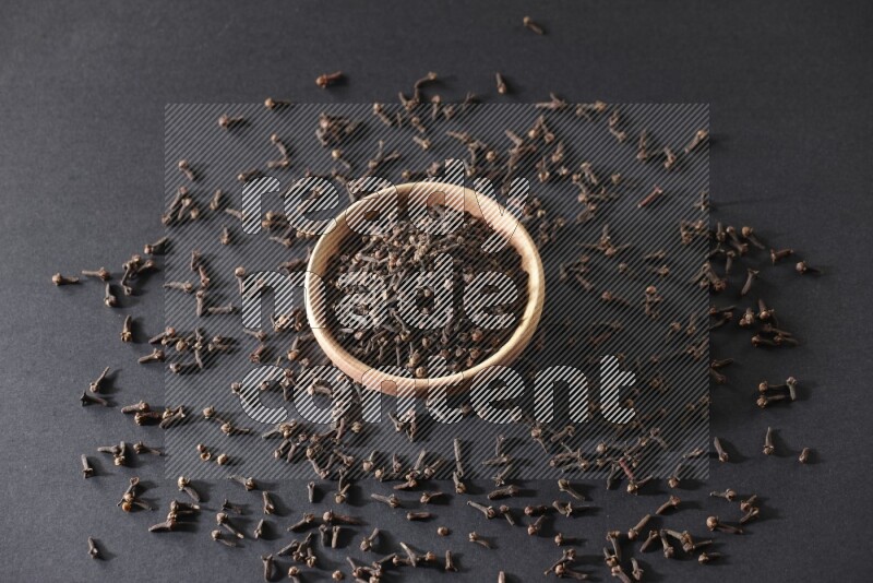 A wooden bowl full of cloves with spreaded whole cloves on a black flooring